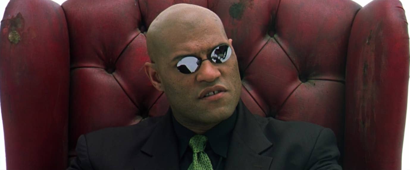 Small mirrored oval sunglasses worn with a dark tailored outfit in a cyberpunk film costume reference.