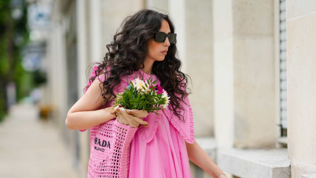 Gabriella Berdugo wears brown square sunglasses Valentino, a pink pleated / gathered dress with maxi Peter Pan ruffled collar from Filkk, a pink crochet grocery beach bag from Prada containing colored flowers, black platform mules, an ankle bracelet.