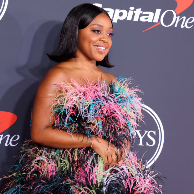 Quinta Brunson, a black woman with a short black bob-style hair cut poses on the red carpet in a multi-colored strapless feather mini dress.