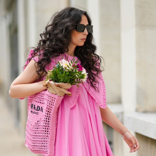 Gabriella Berdugo wears brown square sunglasses Valentino, a pink pleated / gathered dress with maxi Peter Pan ruffled collar from Filkk, a pink crochet grocery beach bag from Prada containing colored flowers, black platform mules, an ankle bracelet.