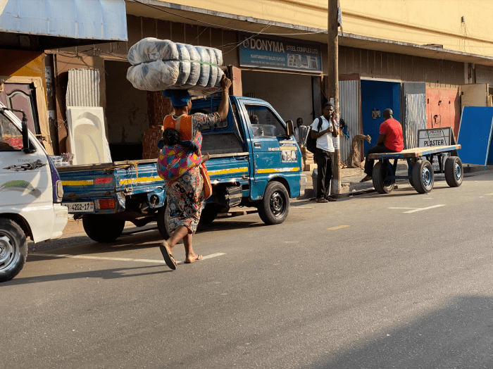 A kayayo carrying a bale of secondhand clothing on her head and a baby on her back through Accra.