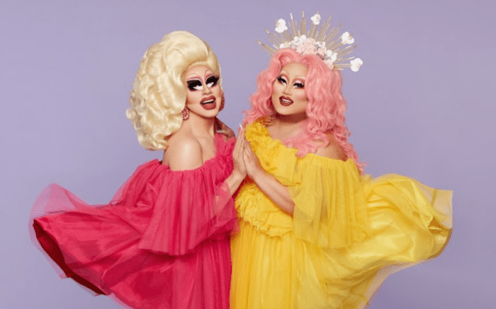 Trixie Mattel, who has blonde hair, wears a floaty.  pink tulle dress, while Kim Chi, wearing pink hair and a headpiece, is wearing a flowing yellow tulle dress.  The two hold hands as they smile for the camera.