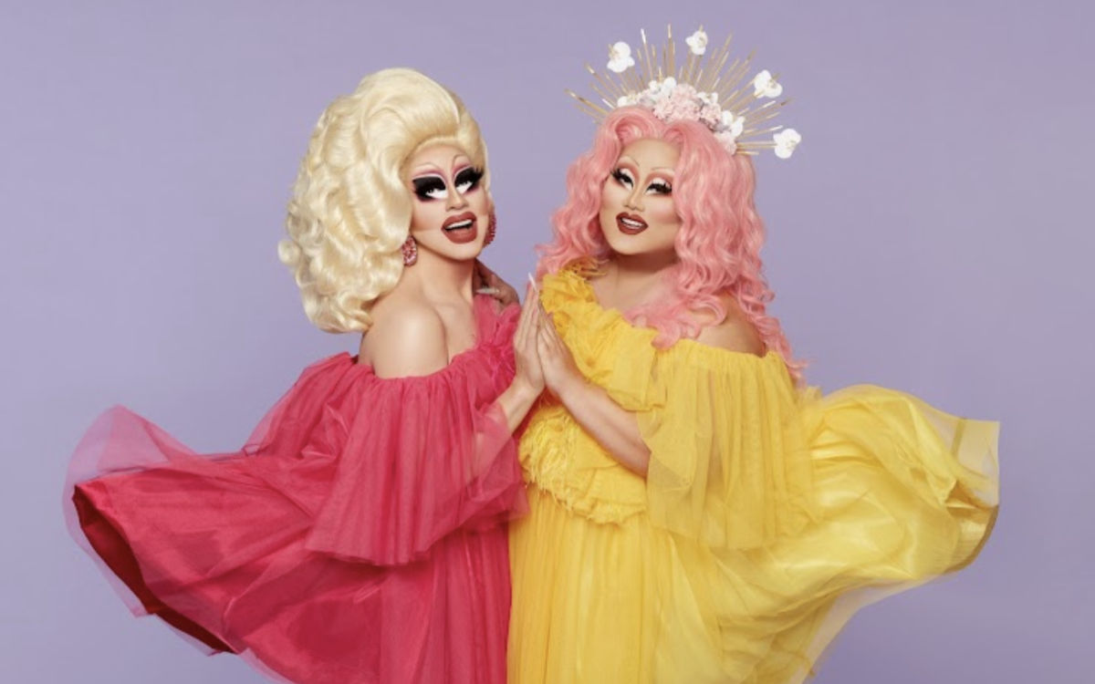 Trixie Mattel, who has blonde hair wears a flowing. pink tulle dress, while Kim Chi, who has pink hair and a headpiece on, wears a flowing yellow tulle dress. The two are holding hands as they smile at the camera.